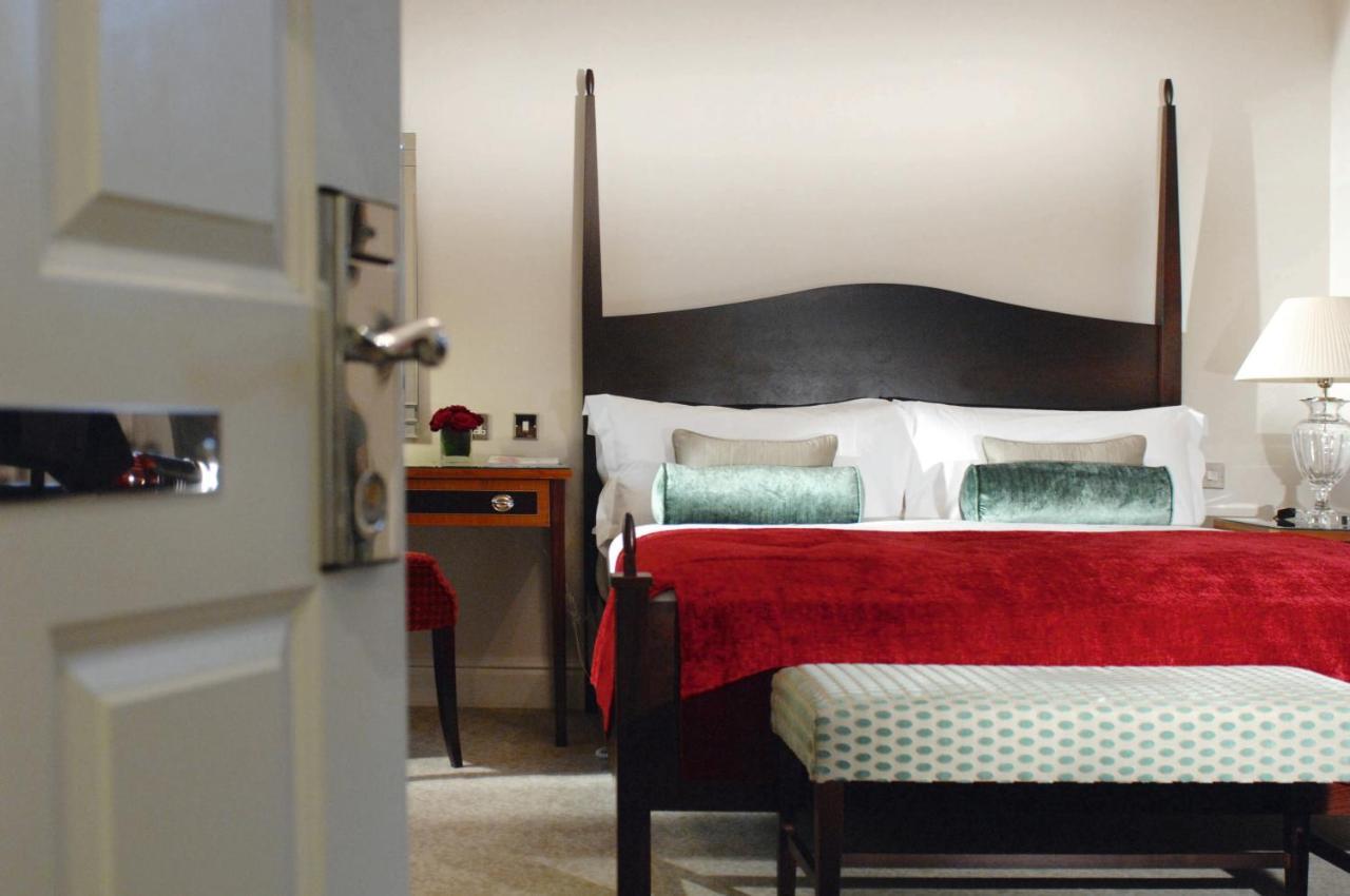 Macdonald Bath Spa Hotel Habitación foto The photo shows a well-decorated hotel room. In the foreground, there is a partially opened door leading into the room. The focal point of the room is a large bed with a dark wooden headboard and stylish red and white bedding, accented with green pil
