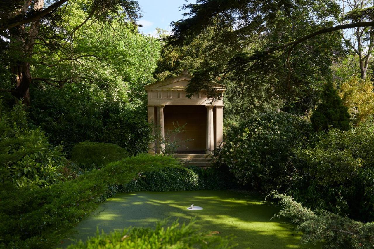 Macdonald Bath Spa Hotel Exterior foto The photo shows a serene garden scene featuring lush greenery and a tranquil pond. In the background, there is a classical-looking structure with columns, resembling a small temple or pavilion. The pond appears to have vegetation covering its surface