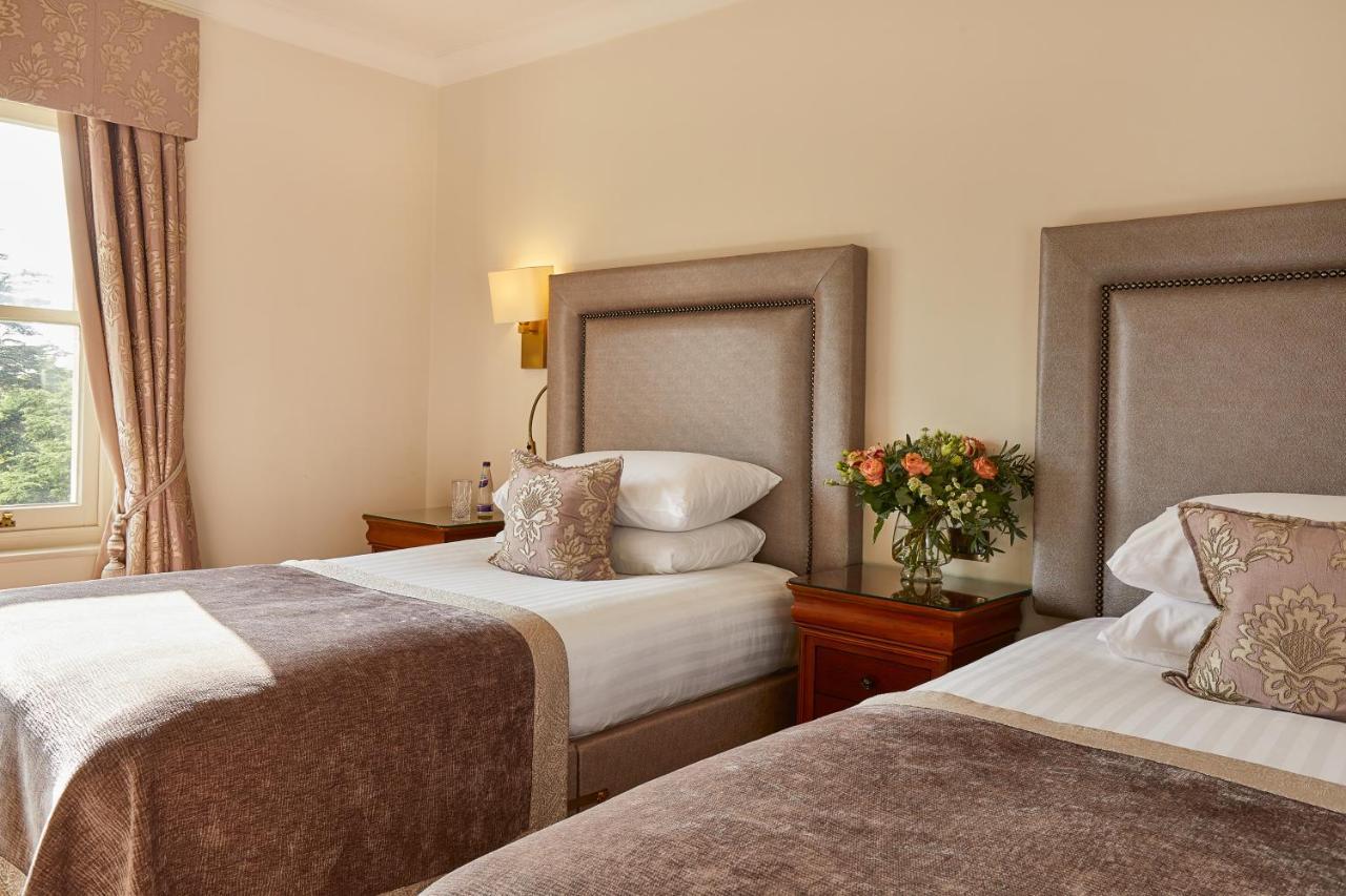Macdonald Bath Spa Hotel Exterior foto The photo shows a hotel room featuring two beds with elegant headboards. Each bed is neatly made with white linens and a soft, decorative throw blanket. There are matching pillows on the beds, and a small, stylish bouquet of flowers is placed on a wo