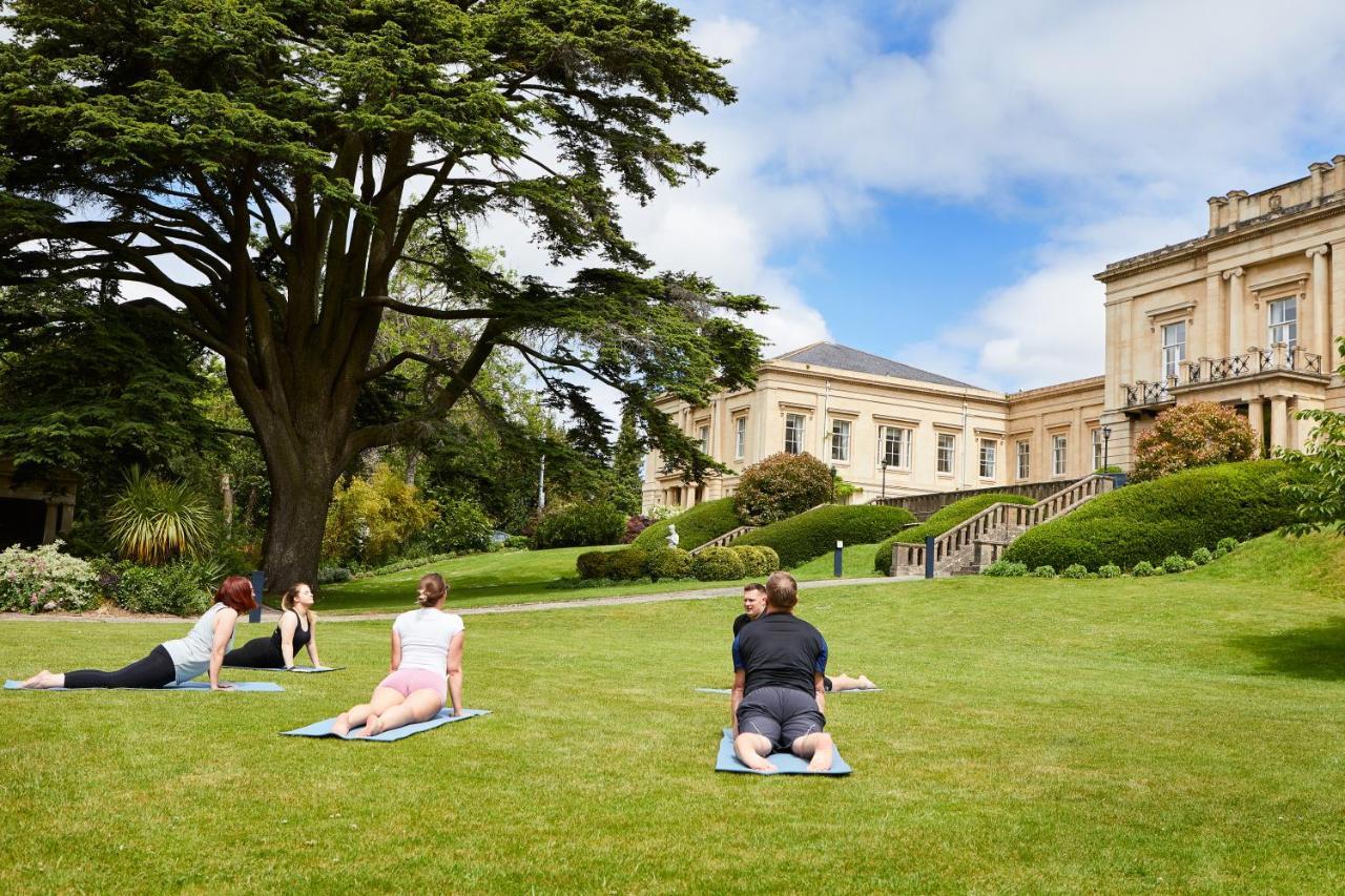 Macdonald Bath Spa Hotel Exterior foto The photo shows a group of people engaging in a yoga session outdoors. They are positioned on yoga mats on a lush green lawn in front of a large, elegant building with columns. Behind them, there are trees and well-maintained gardens, contributing to