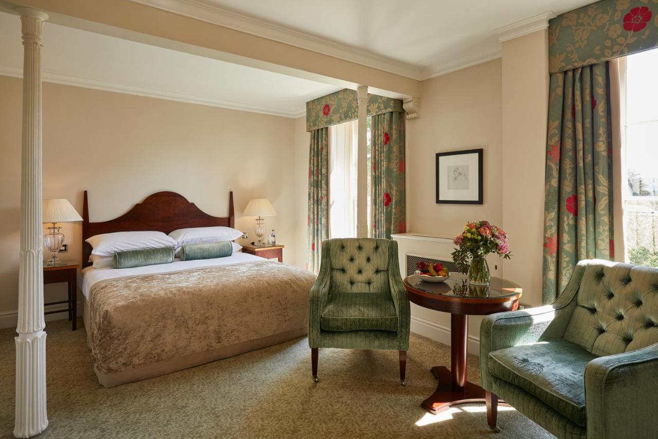 Macdonald Bath Spa Hotel Exterior foto The photo shows a cozy hotel room featuring a large bed with a beige bedspread. There are two green armchairs positioned next to a small round table. The decor includes floral curtains and a neutral color palette on the walls. Soft lighting is provid